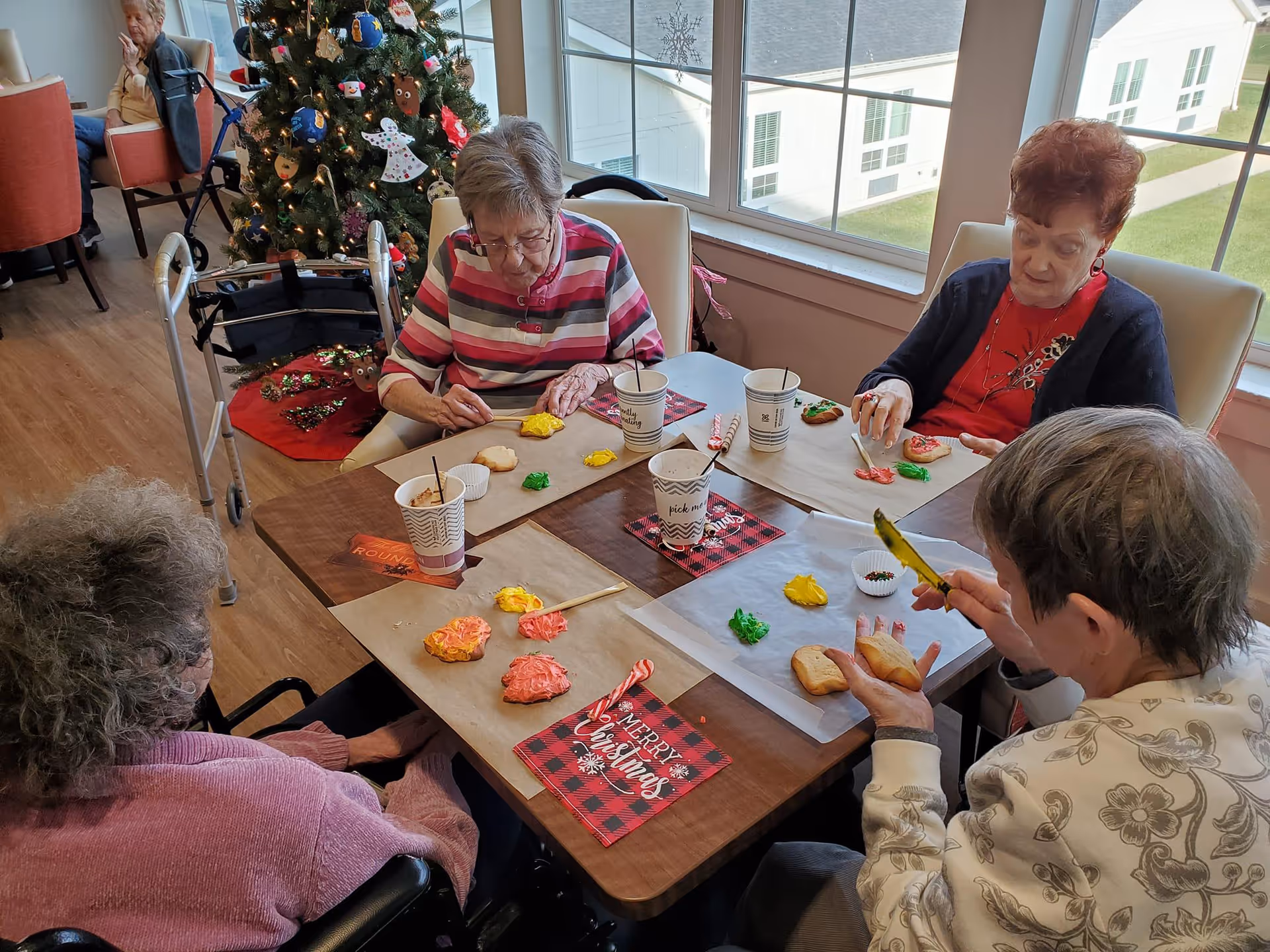 Four elderly women sitting around a table in a well-lit room decorated for Christmas, decorating cookies with colorful icing. A Christmas tree with ornaments is visible in the background near a window showing the outside of the building.