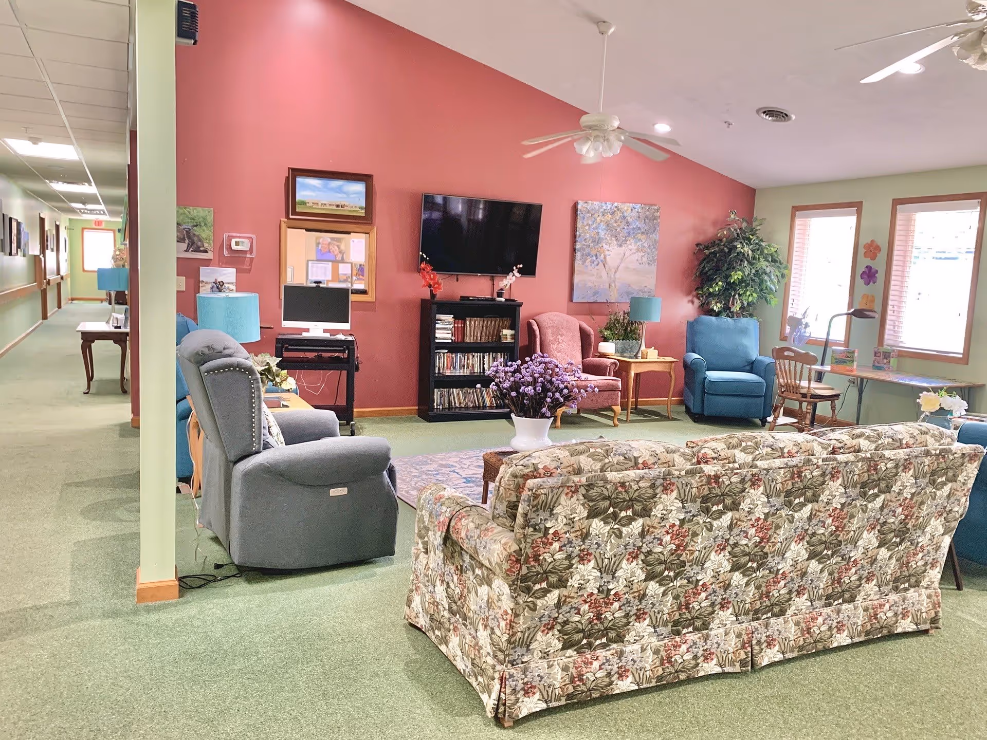 A bright communal living room with a floral sofa, recliners, TV, bookshelf, and seating areas against a pink accent wall.