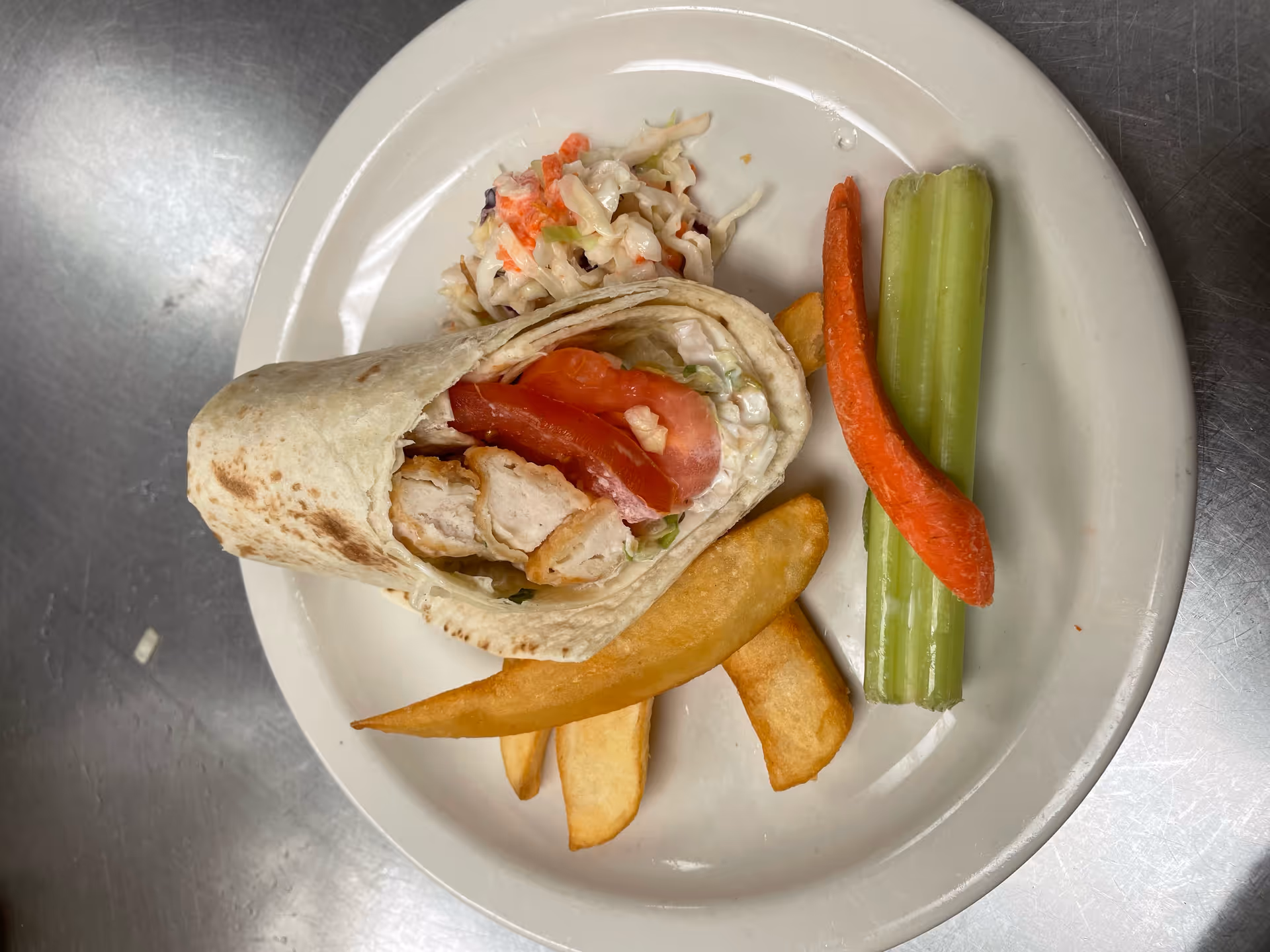 A plate with a chicken wrap containing grilled chicken and tomato slices, served with thick-cut fries, coleslaw, a celery stick, and a carrot stick on a stainless steel surface.
