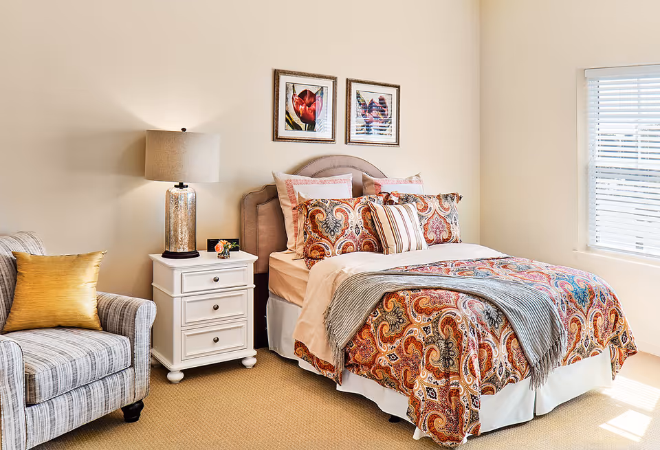 A cozy bedroom featuring a bed with a colorful paisley-patterned comforter and multiple pillows. Next to the bed is a white nightstand with three drawers, a decorative lamp, and a small flower arrangement. A striped armchair with a gold cushion is positioned to the left of the nightstand. Two framed floral artworks hang above the bed, and a window with white blinds allows natural light to brighten the room.