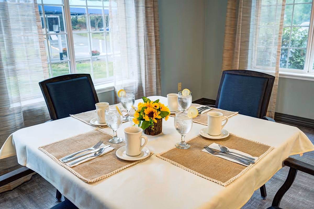 A dining table set for four with beige placemats, white cups and saucers, glasses of water with lemon slices, and a small centerpiece of yellow flowers. The table is covered with a white tablecloth and is positioned near windows with light curtains, allowing natural light into the room.
