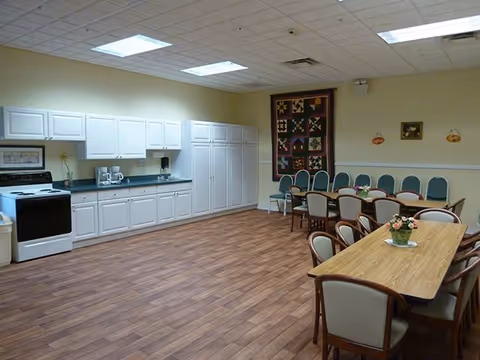 A spacious room with a kitchen area featuring white cabinets, a stove, a coffee maker, and a microwave. The room has wooden flooring and several tables with chairs arranged for dining. A colorful quilt hangs on the wall, and the ceiling has recessed lighting.