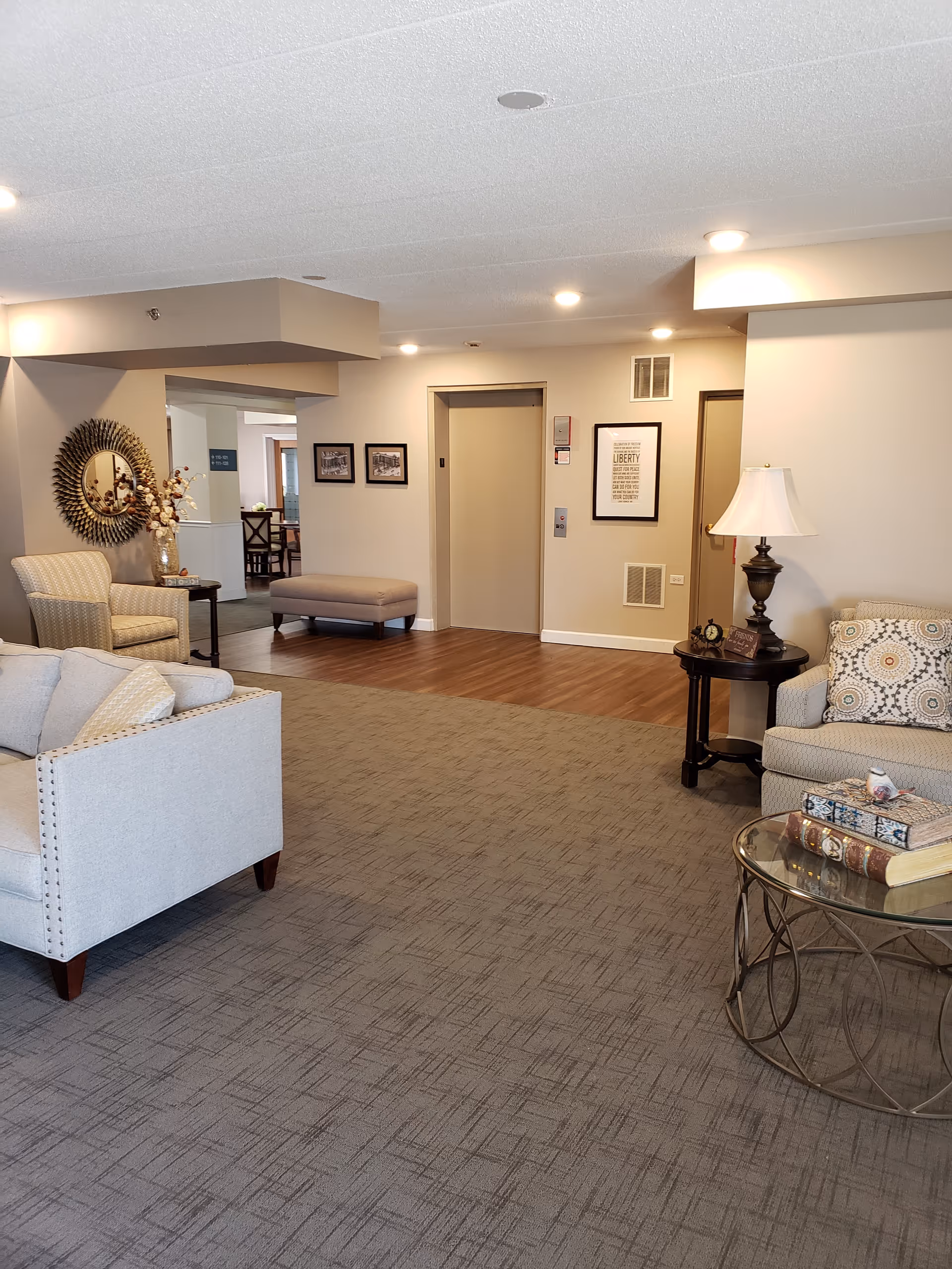 Furnished senior living common area with armchairs, side tables, decorative accents and an elevator at the far wall.