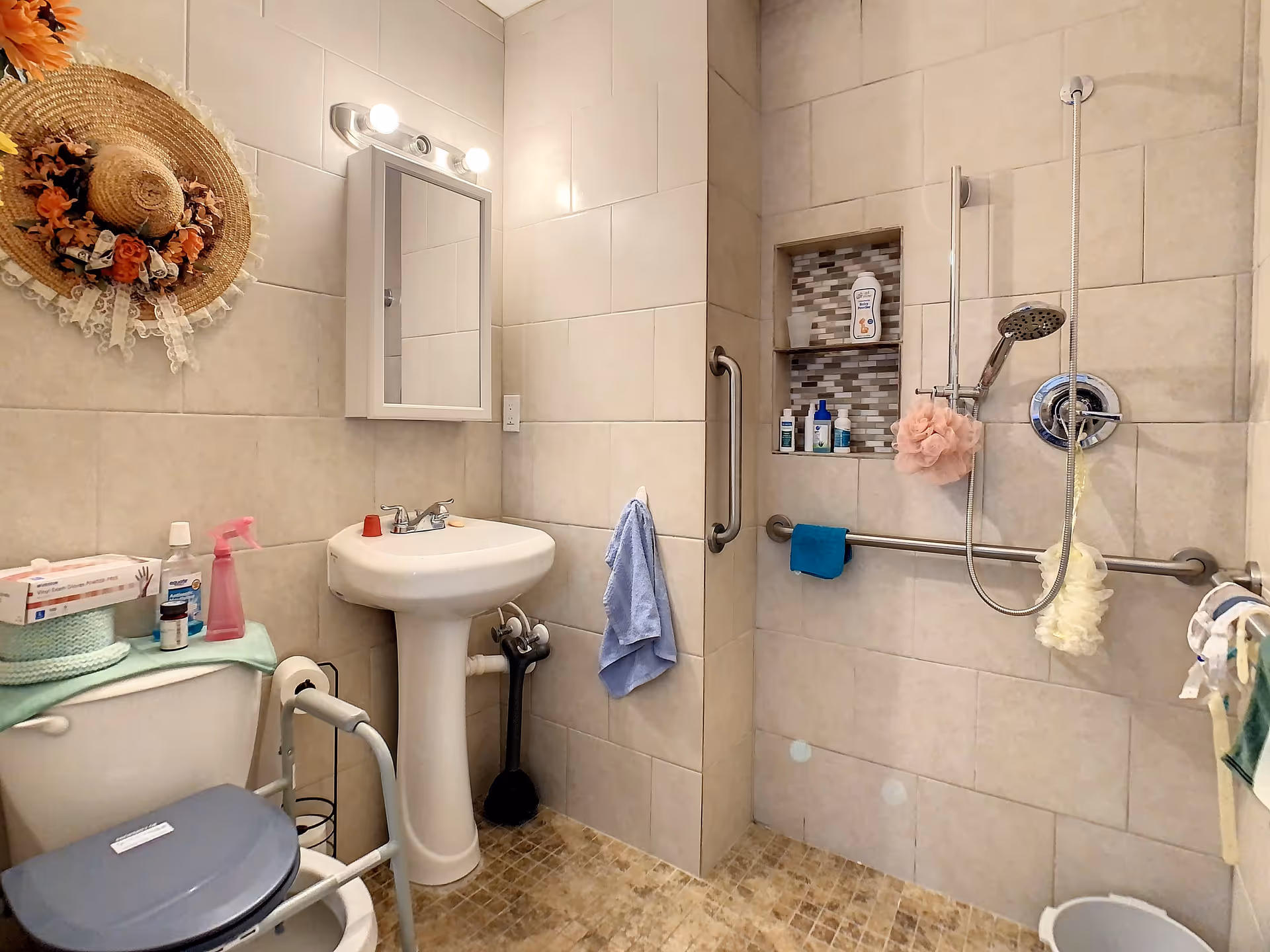 A small, accessible bathroom with beige tiled walls and floor. It features a white toilet with a gray seat and a safety frame, a pedestal sink with a mirror cabinet above it, and a walk-in shower area with a handheld showerhead and grab bars. Various toiletries and cleaning supplies are placed on the toilet tank and in a recessed shelf in the shower. A decorative straw hat with flowers hangs on the wall.