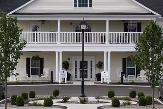 Front entrance of a two-story senior living building with a covered porch, white columns, rocking chairs, a central lamppost, and landscaped circular driveway.