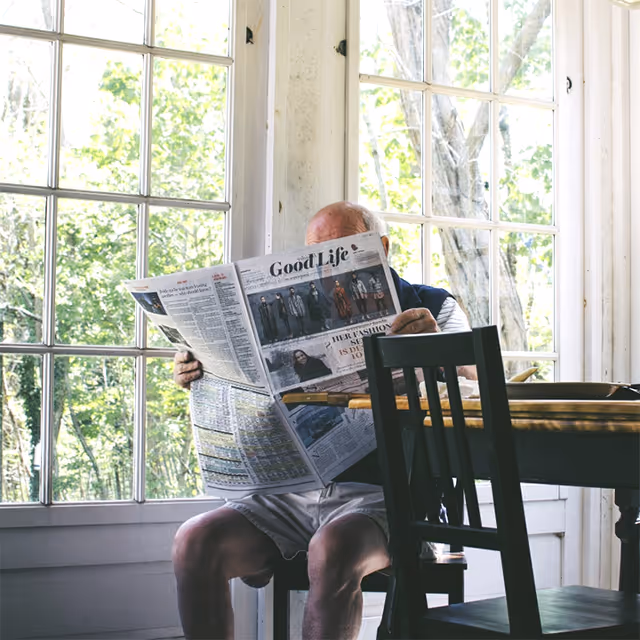 An elderly man sitting at a wooden dining table in a bright room with large windows, reading a newspaper titled 'Good Life'. The room is filled with natural light and shows green trees outside.