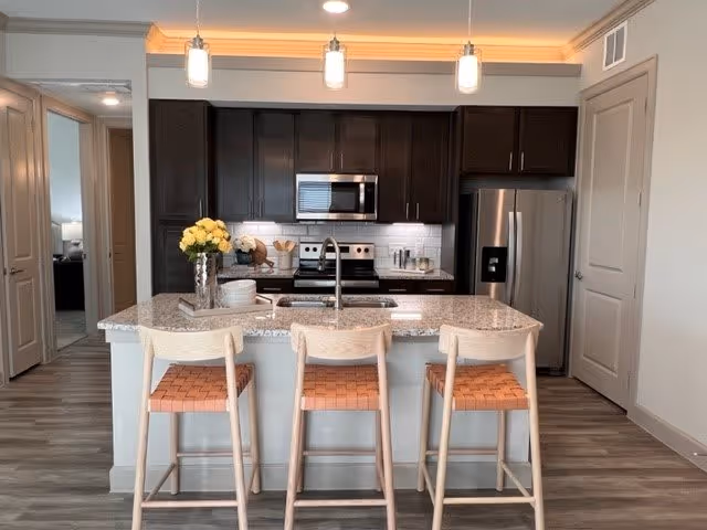 Modern kitchen with a granite countertop island featuring three wooden bar stools with woven seats. The kitchen has dark brown cabinets, a stainless steel refrigerator, microwave, and stove. There are pendant lights hanging above the island and a vase with yellow flowers on the countertop.