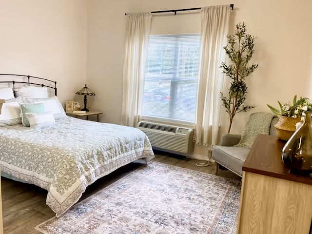 A bright bedroom with a bed covered in a floral quilt, a bedside table with a lamp and picture frames, a window with white curtains, an air conditioning unit below the window, a gray armchair with a green throw, a tall potted plant, a wooden dresser with decorative vases, and a patterned area rug on the wooden floor.