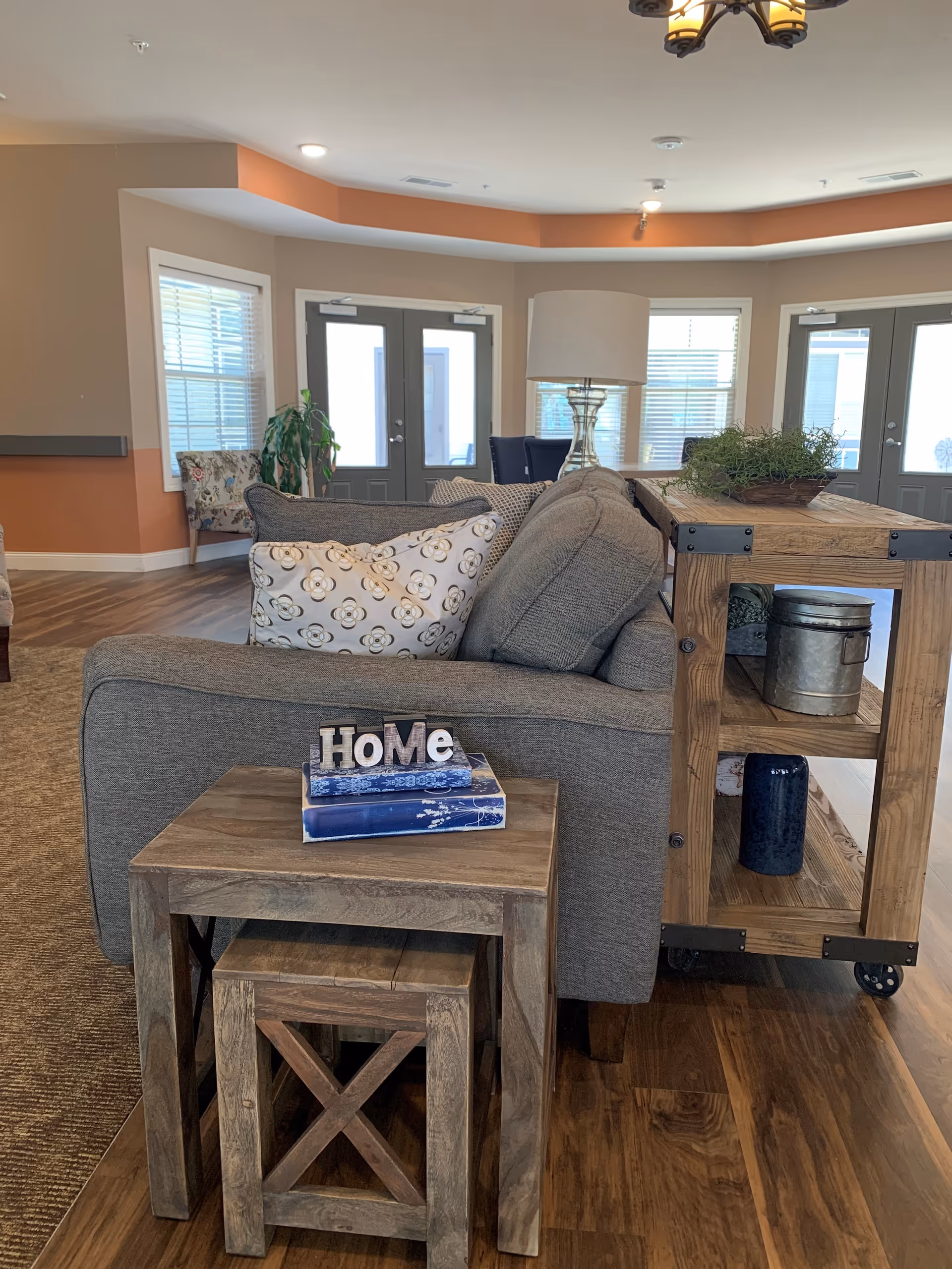 A cozy living room area with a gray couch adorned with patterned pillows. In front of the couch is a small wooden table with a smaller stool underneath, topped with two books and decorative letters spelling 'HoMe'. Behind the couch is a wooden shelving unit with decorative items including a plant, a metal container, and a blue vase. The room has wooden flooring, beige and orange walls, multiple windows with blinds, and glass doors leading outside. A ceiling light fixture is visible above.