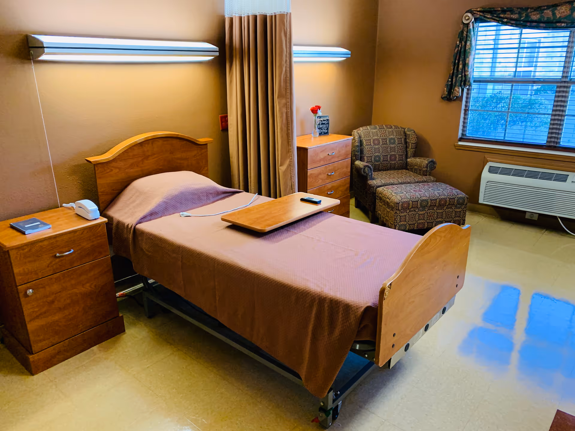 A single hospital-style bed with a wooden headboard and footboard, covered with a brown blanket, in a room with beige walls and a tiled floor. Next to the bed is a wooden nightstand with a telephone and a book. A wooden overbed table with a remote control rests on the bed. In the corner, there is a patterned armchair with an ottoman, a wooden dresser with a small vase holding a red flower, and a window with blinds and floral curtains. A wall-mounted air conditioning unit is below the window.