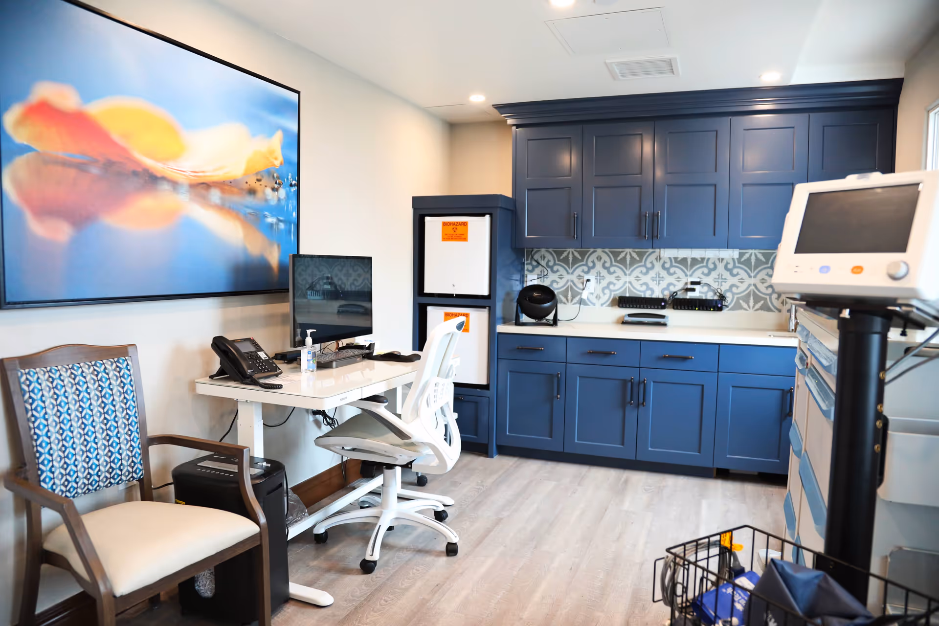 A clean and modern medical office room with blue cabinets, a white desk with a computer monitor, telephone, and hand sanitizer, a white office chair, a wooden chair with patterned upholstery, and medical equipment including a monitor and biohazard disposal containers.