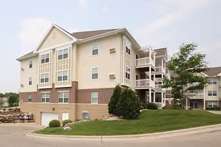 Three-story beige senior apartment building with balconies, garage entrances, and a small landscaped lawn under a partly cloudy sky.