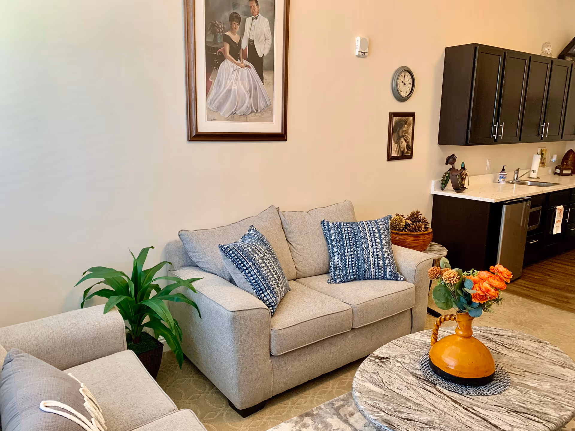 A cozy living room area with a light gray loveseat and armchair, both adorned with blue patterned throw pillows. A round marble coffee table holds an orange vase with orange and green flowers. Behind the loveseat, there is a framed portrait of a couple dressed formally, a wall clock, and a smaller framed picture. To the right, a kitchen area with dark cabinets, a countertop, a sink, and various items is visible. A green potted plant is placed beside the armchair.