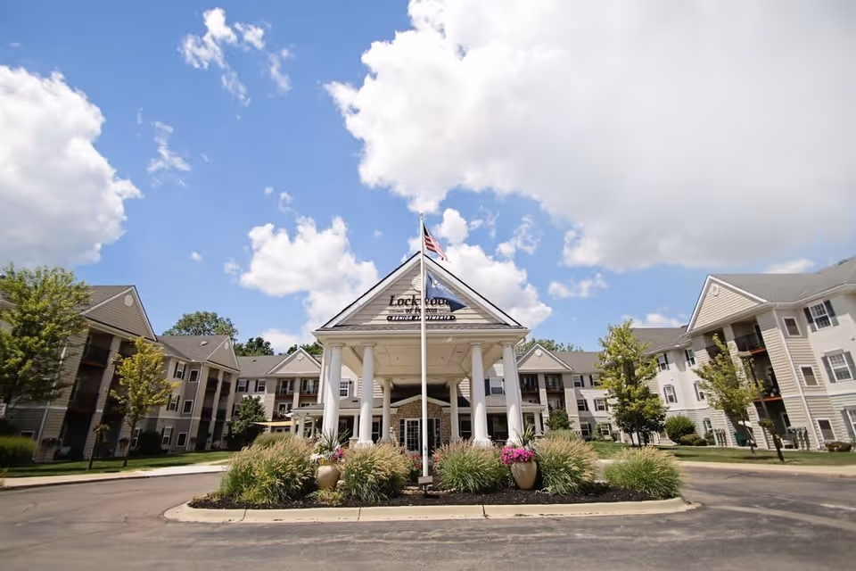 Front exterior view of Lockwood of Fenton senior living facility with a central entrance featuring white columns, an American flag on a flagpole, and surrounding landscaped greenery under a partly cloudy sky.