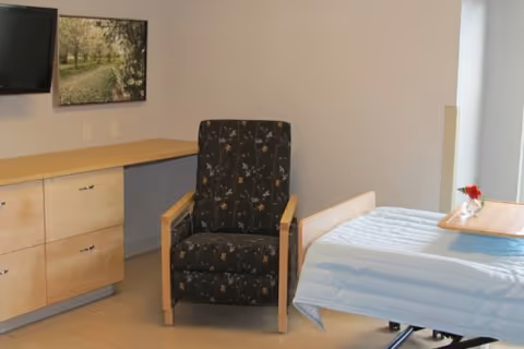 A senior living facility room featuring a hospital-style bed with a light blue blanket, a wooden overbed table with a red flower in a vase, a black armchair with a floral pattern, a wooden cabinet with drawers, a mounted flat-screen TV, and a framed picture of a tree-lined path on the wall.