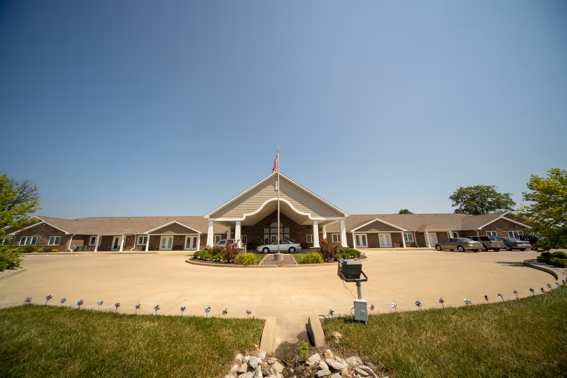 Wide exterior view of a single-story senior living facility building with a large covered entrance, an American flag on a flagpole, several parked cars, and a clear blue sky.