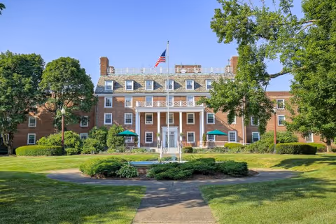 Front exterior view of a large brick building with multiple windows and chimneys, a central entrance with white columns, an American flag on a flagpole in front, surrounded by green trees and well-maintained lawns.