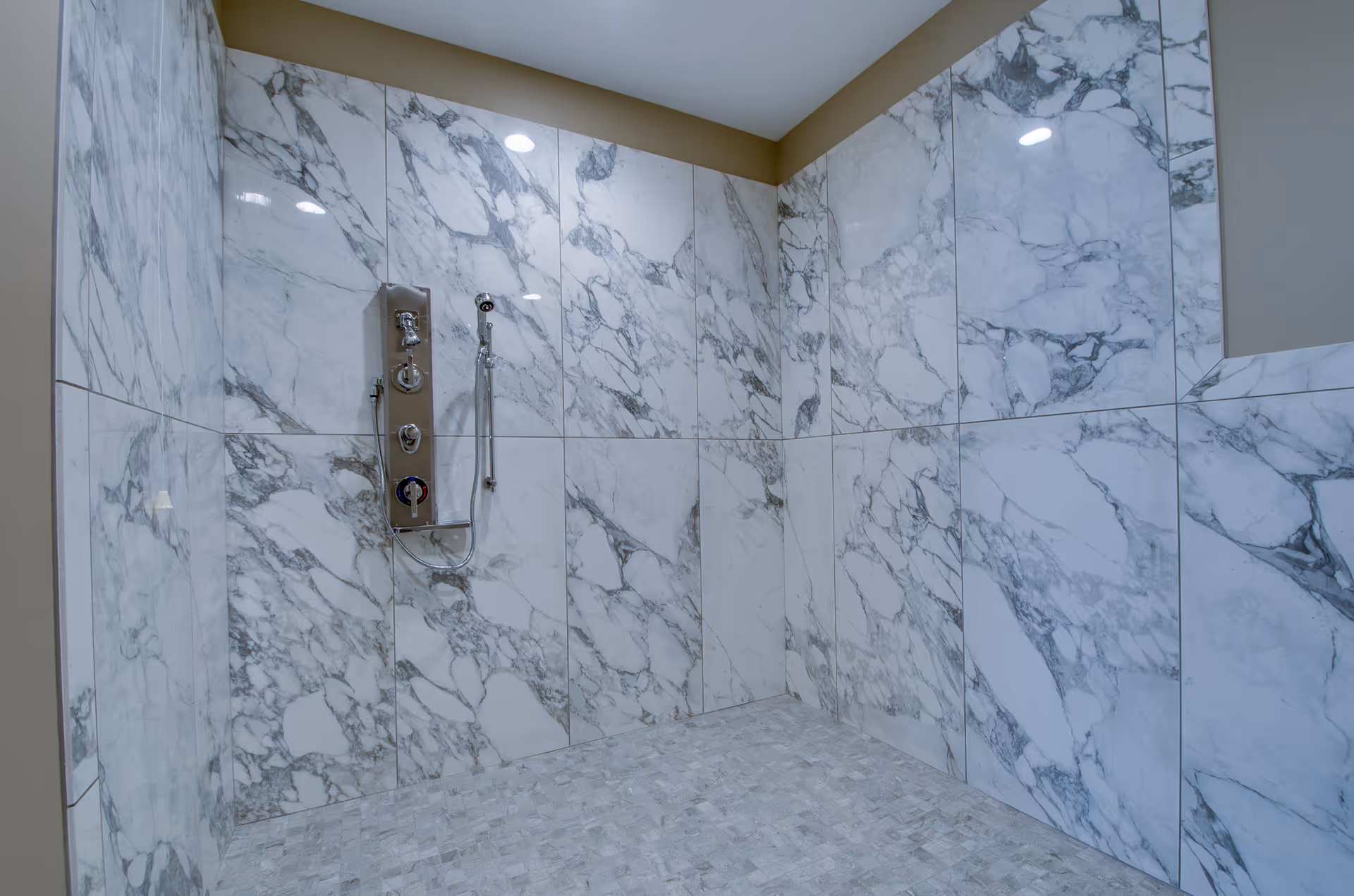 A modern shower area with large white and gray marble tiles covering the walls and smaller matching tiles on the floor. The shower features a sleek metal control panel with knobs and a handheld showerhead attached to the wall.