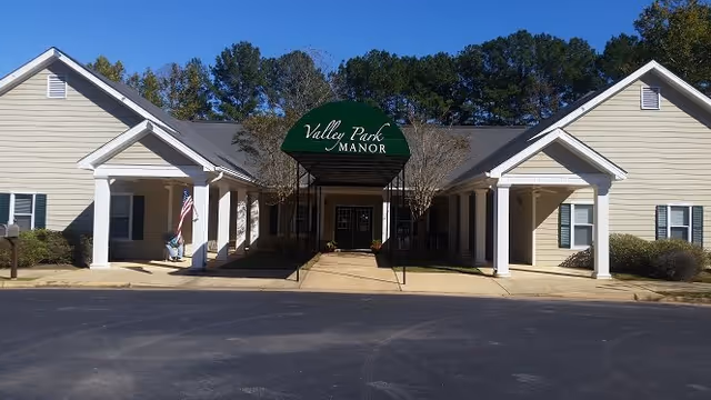 Front exterior view of Valley Park Manor Assisted Living & Memory Care building with a covered entrance featuring a green awning with the facility name. The building is light-colored with white trim and surrounded by trees under a clear blue sky.