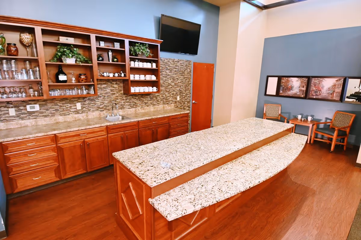 Interior view of a kitchen area with wooden cabinets, a granite countertop island, open shelves holding glassware and mugs, a small sink, a wall-mounted TV, and a seating area with two chairs and a small table against a blue accent wall with framed artwork.