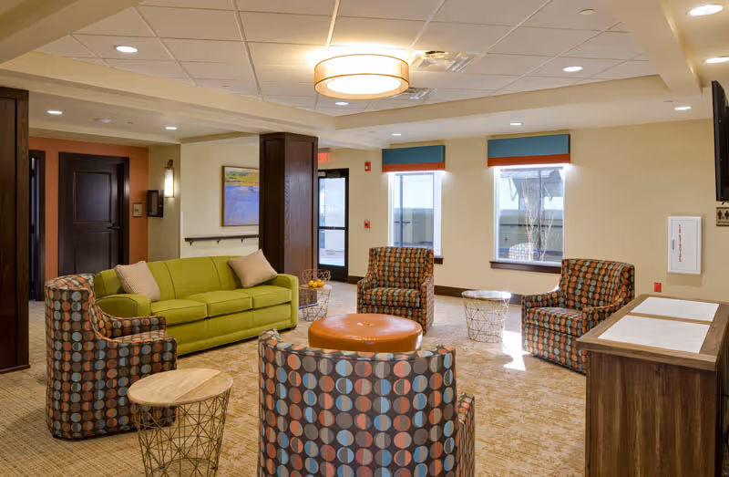 A cozy senior living facility common area with a green sofa, four patterned armchairs, a round orange ottoman, and small round side tables. The room has beige walls, carpeted floor, two windows with blue and orange valances, and a ceiling light fixture. There is a dark wooden cabinet on the right and a door leading outside in the background.