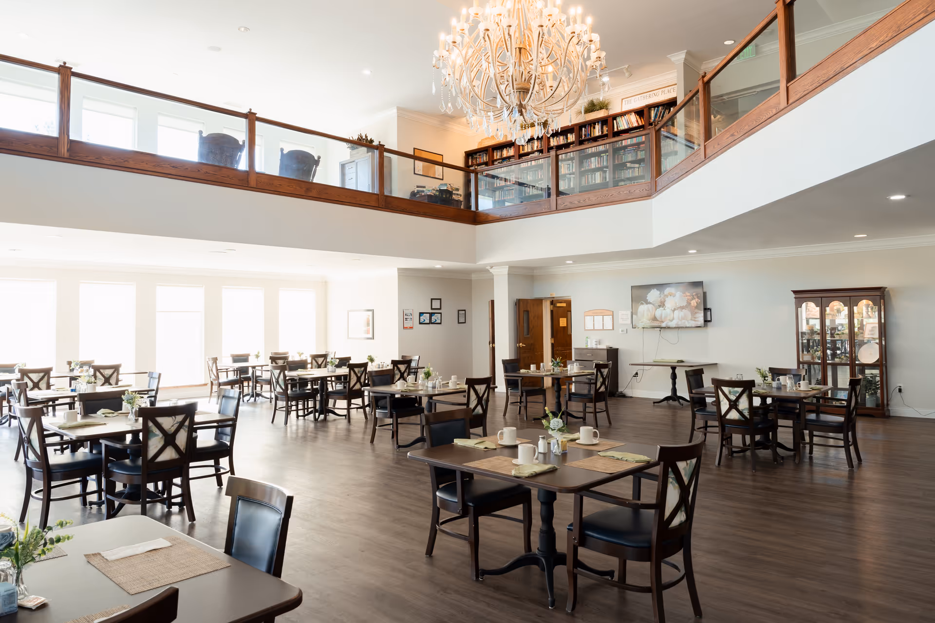 A spacious and well-lit dining room with multiple wooden tables and chairs arranged neatly. Each table is set with placemats, napkins, cups, and small flower vases. The room features large windows allowing natural light to flood in, a chandelier hanging from the ceiling, and a second-floor balcony with bookshelves and seating visible above. There is a television mounted on the wall and a glass cabinet displaying decorative items.