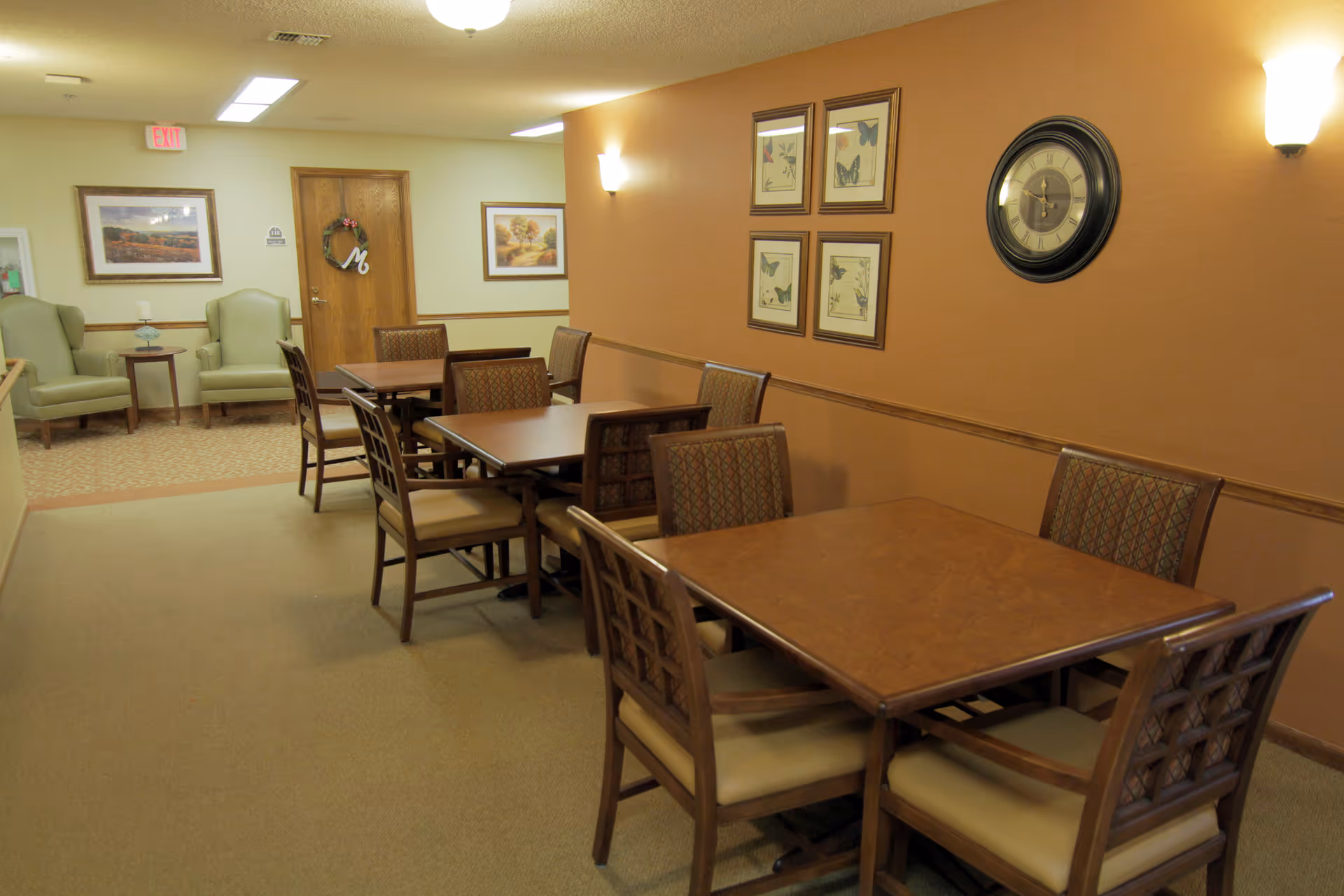 Dining room with multiple wooden tables and chairs, a wall clock and framed artwork on warm-colored walls.