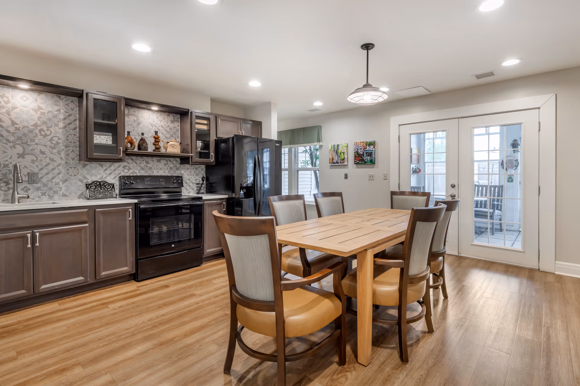 A modern kitchen and dining area with wooden flooring, dark brown cabinets, a black refrigerator and stove, a wooden dining table with six chairs, and glass double doors leading to an outdoor space.