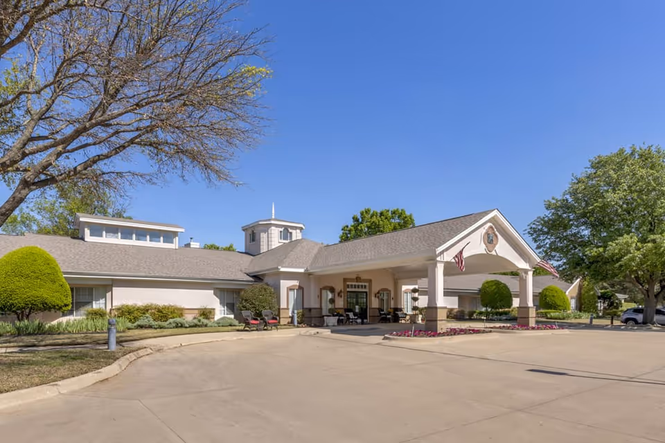 Exterior view of Brookdale Stonebridge Ranch senior living facility showing a large building with a covered entrance, manicured bushes, trees, and a clear blue sky.