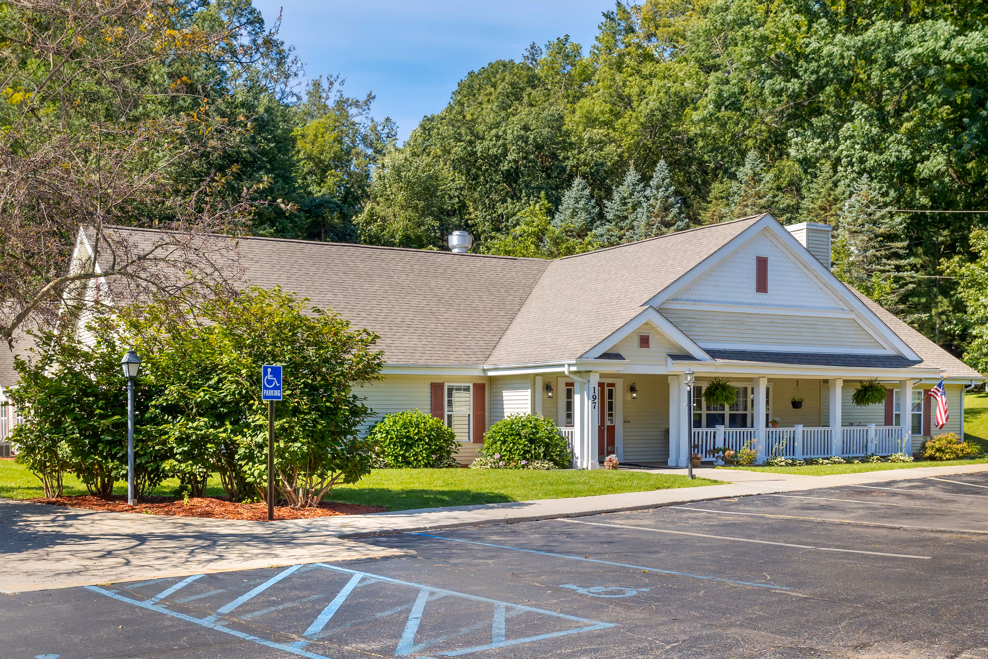 Exterior view of a single-story building with a beige roof and light-colored siding, surrounded by green trees and bushes. There is a small porch with white railings and hanging plants, an American flag on the right side, and a parking lot with a marked handicapped parking space in the foreground.