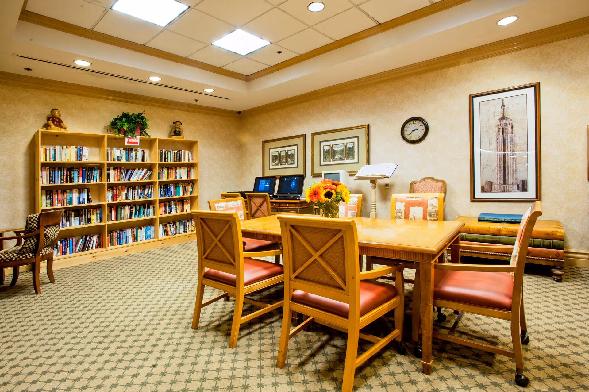 A cozy reading and computer room with a wooden table surrounded by six chairs with red cushions. A vase with sunflowers is placed on the table. In the background, there is a bookshelf filled with books, two desktop computers on a desk, framed pictures on the wall, a clock, and a framed picture of a tall building. The room has patterned carpet and warm lighting.