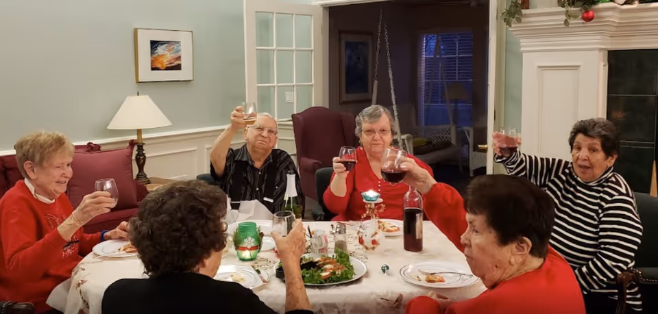 A group of six elderly people sitting around a dining table raising their glasses in a toast. The table is set with plates, a salad, and a bottle of wine. The room has light-colored walls, a lamp, framed artwork, and a fireplace decorated with holiday ornaments.