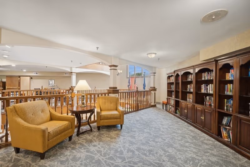 A cozy sitting area in a senior living facility with two mustard yellow armchairs and a round wooden side table with a lamp. The space overlooks a lower level through wooden railings. On the right side, there is a large wooden bookshelf filled with books. The area is well-lit with natural light coming from a large arched window in the background.