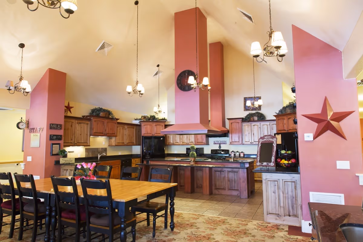 A spacious kitchen and dining area with wooden cabinets, black appliances, and a large wooden dining table surrounded by black chairs. The walls are painted in warm tones with red accent columns, decorated with a large red star and greenery on top of the cabinets. Multiple hanging light fixtures illuminate the room.