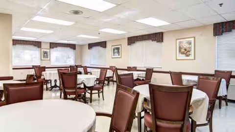 A bright dining hall with round tables, white tablecloths, and red cushioned chairs.
