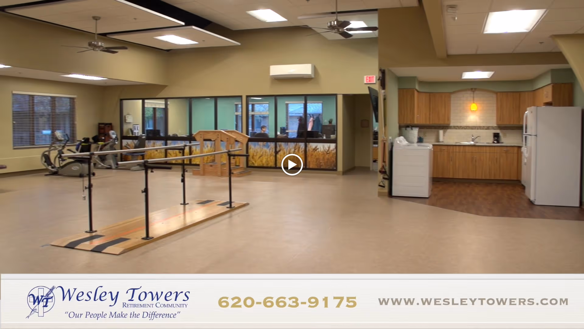 Interior view of a senior living facility room featuring exercise equipment including a stationary bike and parallel bars for physical therapy. There is a kitchenette area with wooden cabinets, a refrigerator, and a washing machine. Large windows and ceiling fans are visible, along with a reception or office area in the background.