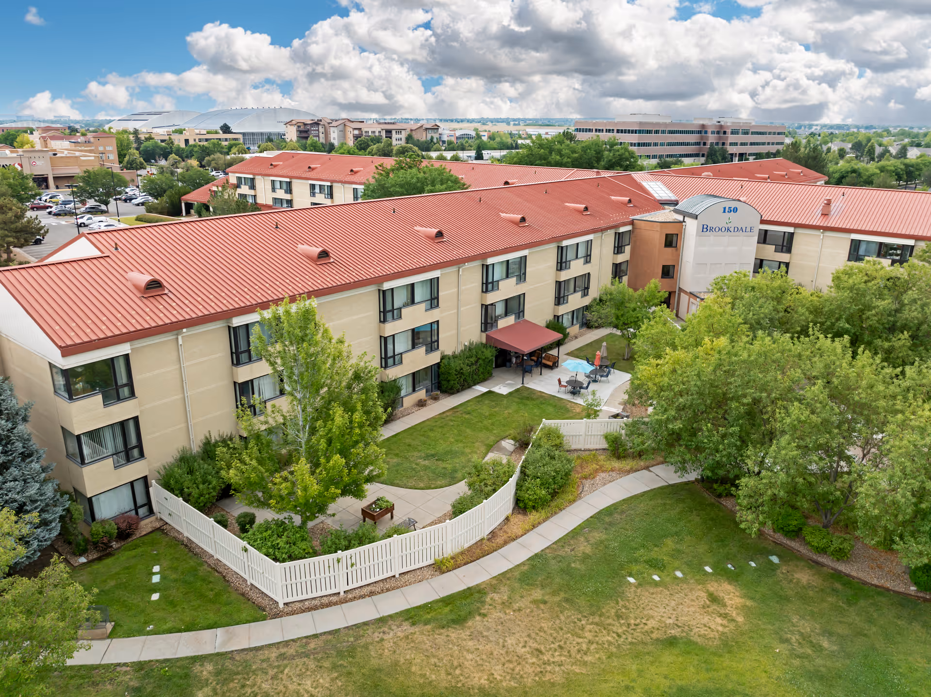 Aerial view of Brookdale Lowry senior living facility showing a large beige building with a red roof, surrounded by green trees and landscaped gardens. There is a fenced patio area with outdoor seating and umbrellas, and a curved walkway through the grass in the foreground. The sky is partly cloudy.