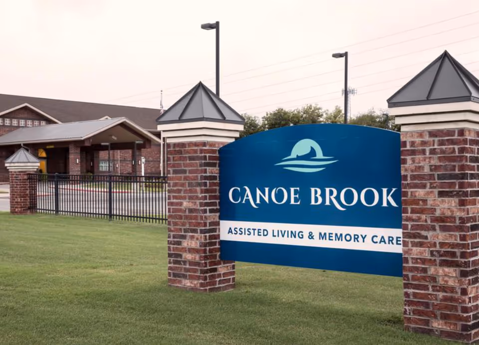 Outdoor view of the entrance sign for Canoe Brook Assisted Living & Memory Care, featuring a blue sign with white text mounted between two brick pillars, with the facility building and a black metal fence visible in the background.