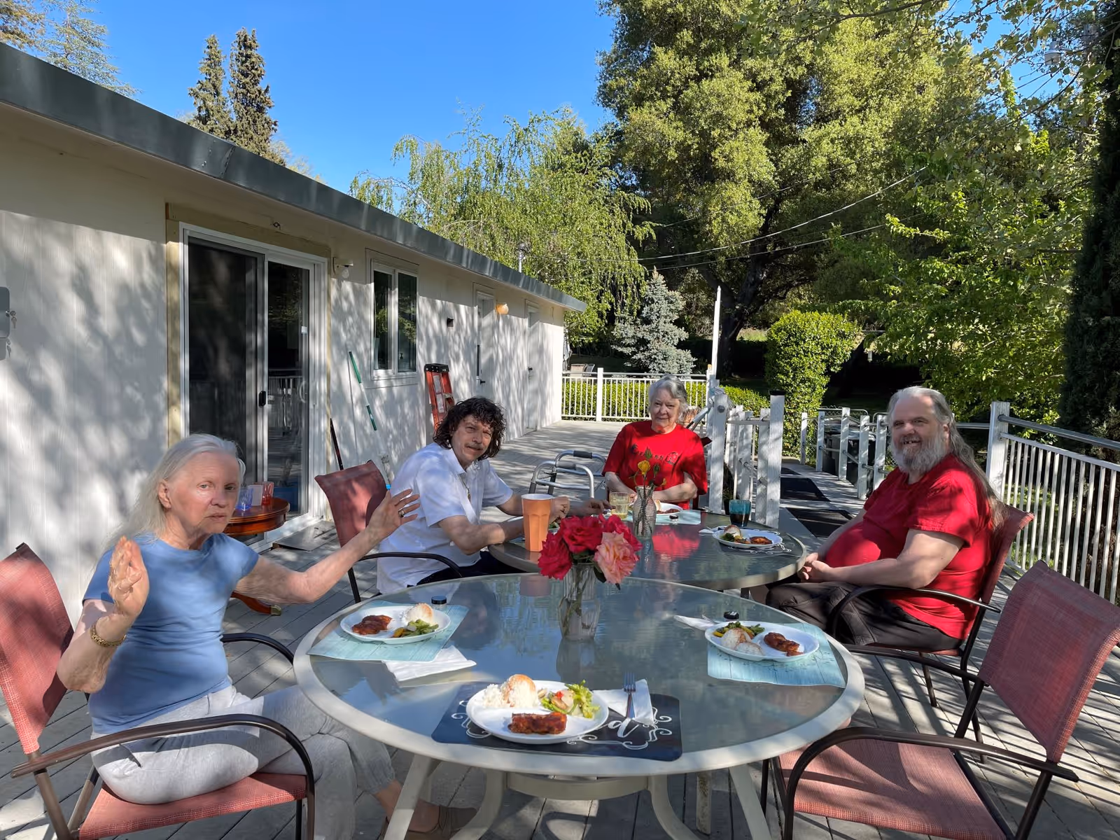 Four elderly people sitting around a glass outdoor table on a patio, enjoying a meal together. The table has plates with food and a vase with flowers. The setting is sunny with trees and greenery in the background, next to a white building.