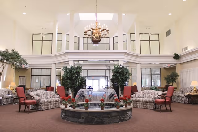 Spacious senior living facility common area with a central stone fountain decorated with red flowers. Surrounding the fountain are floral-patterned sofas and red cushioned chairs arranged in seating groups. The room features high ceilings with large windows, a brass chandelier hanging from above, and several potted plants adding greenery to the space.