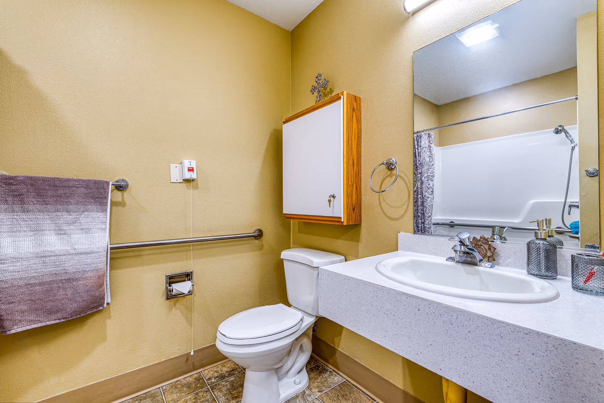A clean bathroom with beige walls featuring a white toilet, a white sink with a speckled countertop, a large mirror above the sink, a wall-mounted cabinet with a decorative cross on top, a towel rack with a striped towel, a grab bar next to the toilet, and a shower with a patterned curtain visible in the mirror.