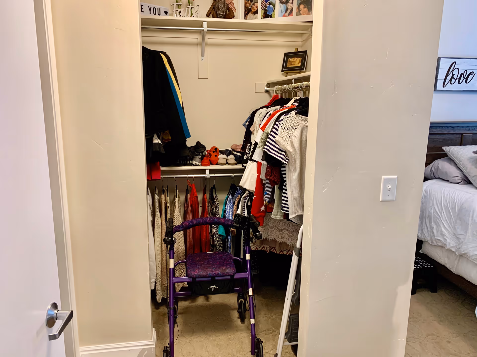 View into a walk-in closet with clothes hanging on rods and shoes on a shelf. A purple walker is positioned in front of the closet. To the right, part of a bedroom with a bed, pillows, and a wall decoration with the word 'love' is visible.