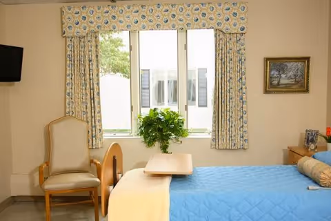 A nursing facility room with a single bed covered in a blue quilt and a beige blanket. There is a wooden overbed table on the bed, a beige cushioned chair next to the bed, and a window with floral curtains letting in natural light. A small green plant is placed on the windowsill, and a framed picture hangs on the wall above a wooden dresser.
