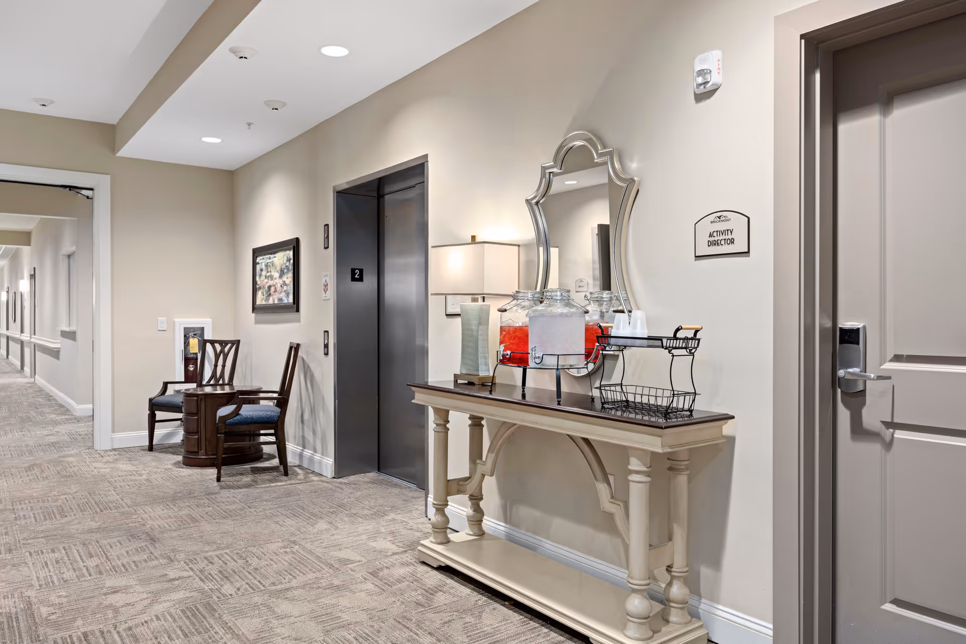 Interior hallway of a senior living facility with an elevator door labeled '2', a decorative console table with water dispensers and cups, a wall mirror, a lamp, and a sign indicating the Activity Director's office. There are two chairs and a small table along the hallway, with neutral-colored walls and carpeted flooring.