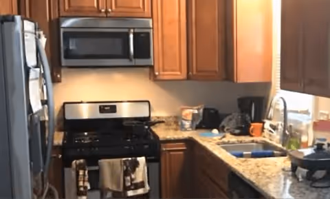 A kitchen with wooden cabinets, a stainless steel microwave above a gas stove with towels hanging on the oven door, a granite countertop with various kitchen items including a coffee maker, and a window above the sink letting in natural light.