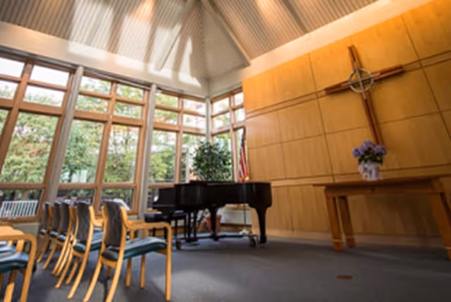 Sunlit chapel-like common room with a grand piano, rows of chairs, large windows, and a wooden wall cross.