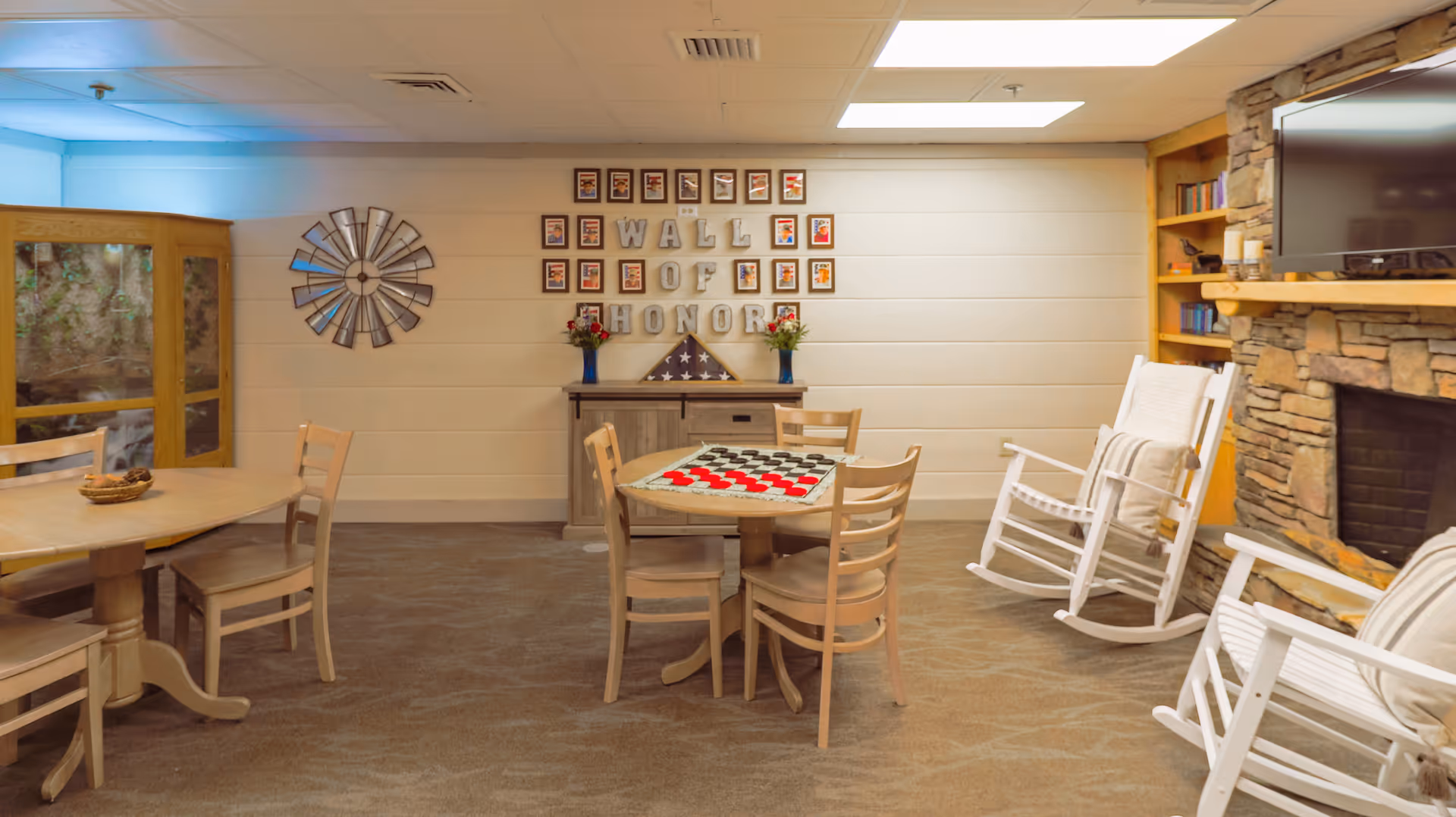 Cozy senior living common room with tables and chairs, rocking chairs by a stone fireplace, and a "Wall of Honor" display.