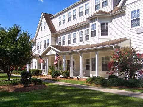 Front exterior of a multi-story white senior living building with a covered porch, American flags, and landscaped lawn.