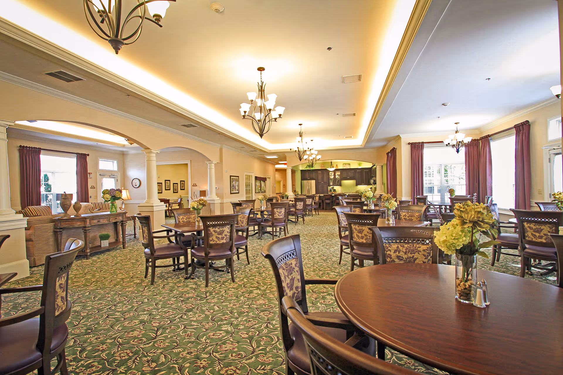 A spacious dining room with multiple round and square wooden tables surrounded by chairs with patterned upholstery. The room features green patterned carpet, cream-colored walls, large windows with red curtains, and elegant chandeliers hanging from the ceiling. There is a kitchen area with dark cabinets and stainless steel appliances visible in the background.