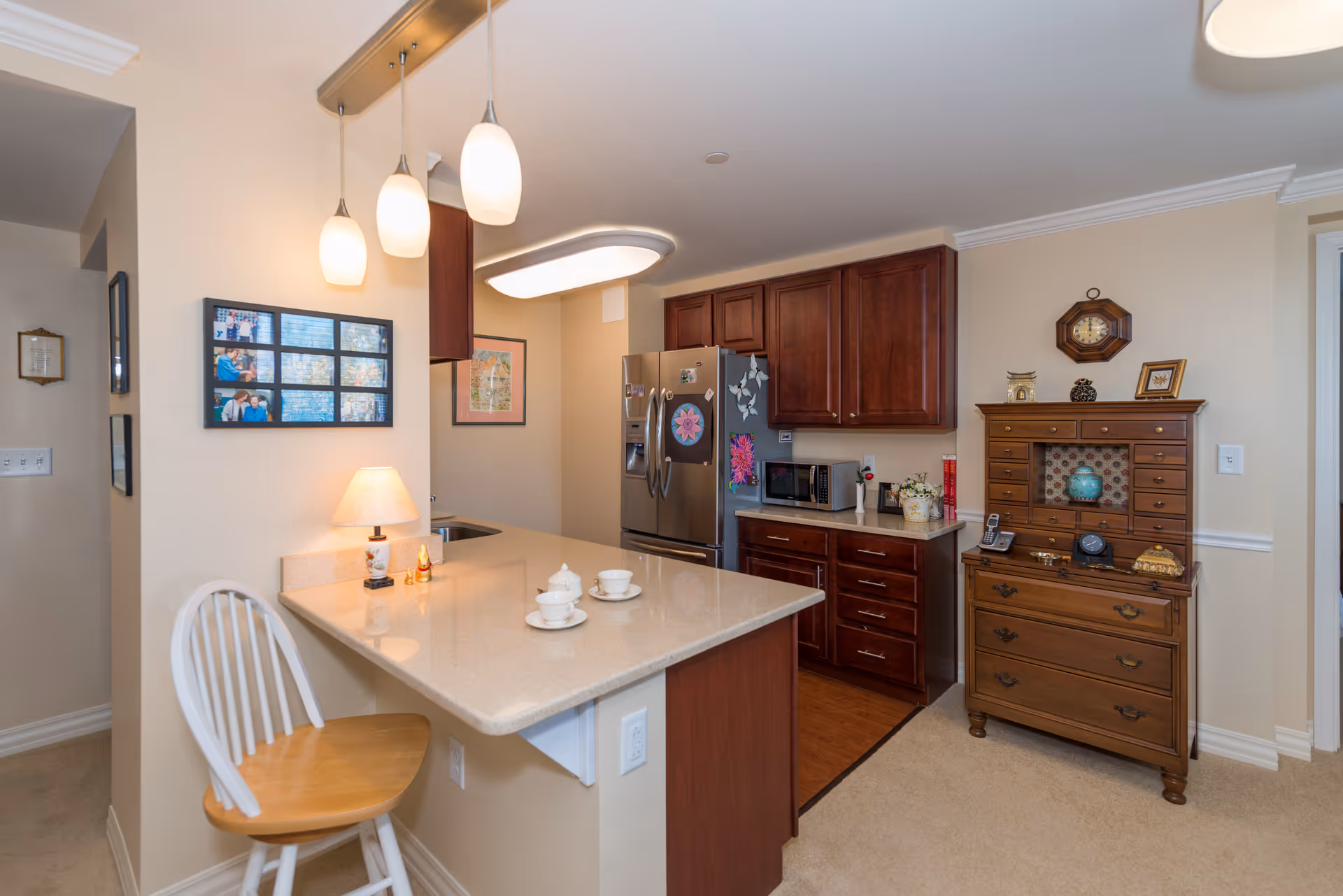 Interior view of a kitchen area with a beige countertop island featuring two teacups and saucers, a wooden chair with a white backrest, and three hanging pendant lights above. The kitchen has dark wooden cabinets, a stainless steel refrigerator decorated with magnets, a microwave, and various small decorative items. Adjacent to the kitchen is a wooden dresser with multiple drawers and decorative items on top, including a clock and framed pictures. The walls are painted light beige, and there is a framed photo collage and artwork on the walls.